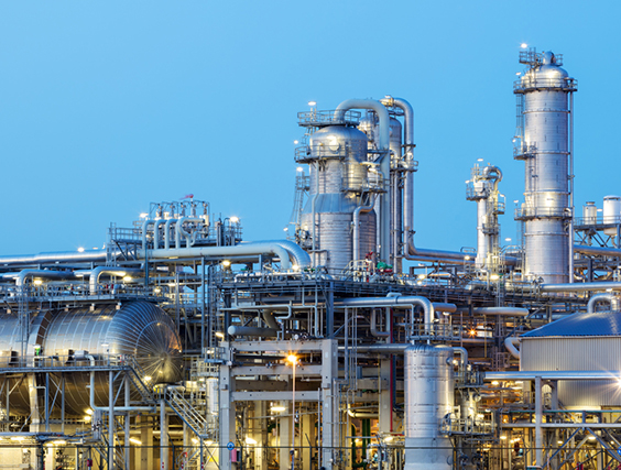 A modern, illuminated refinery is visible at dusk. Lights from the buildings and streets glow. Several distillation towers can be seen as well as glowing moon. Long exposure with tripod.